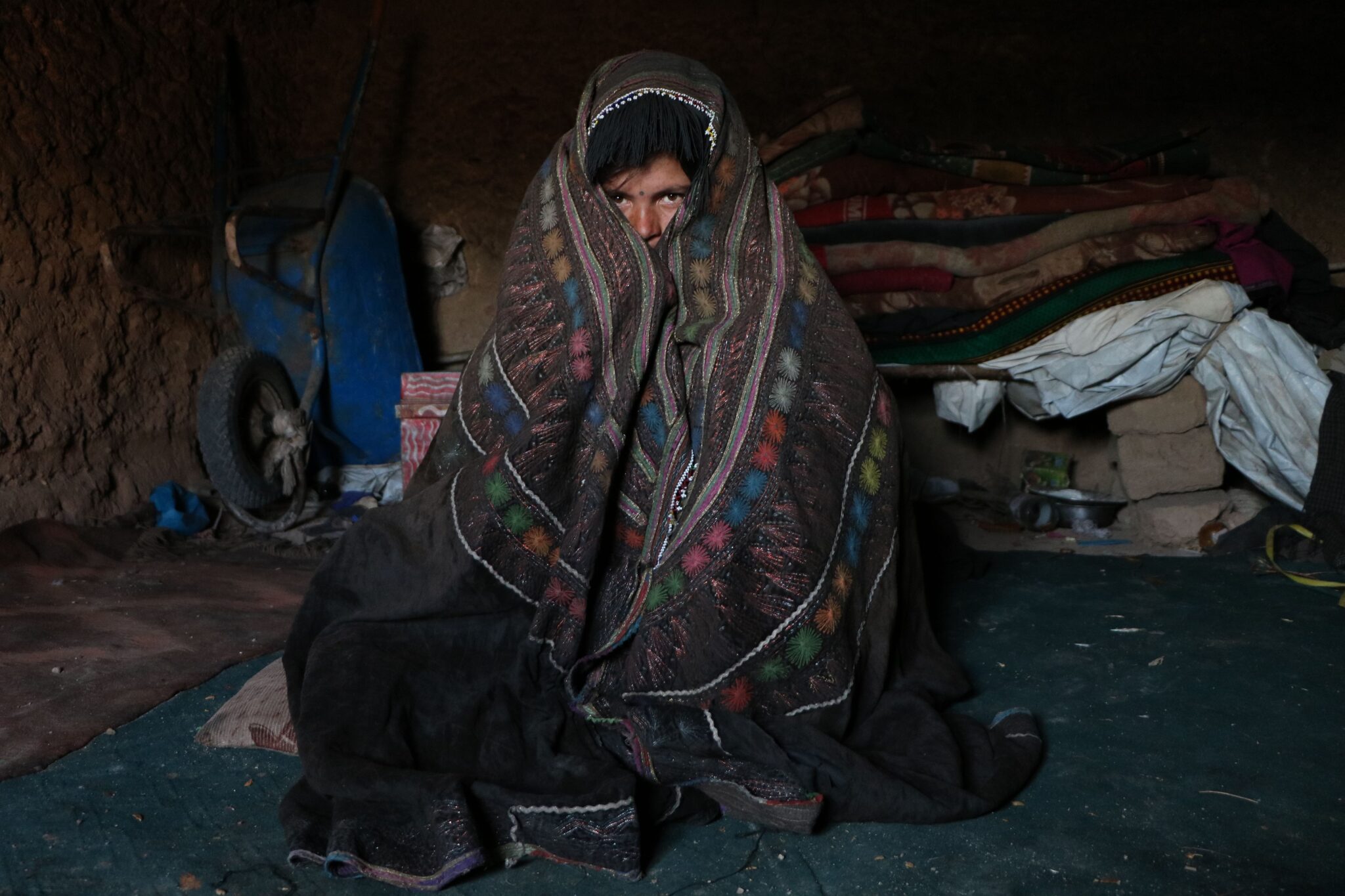A person wrapped in a colorful blanket sitting in a dimly lit indoor space in Herat, Afghanistan. Photo by Morteza Rezaiy, pixel