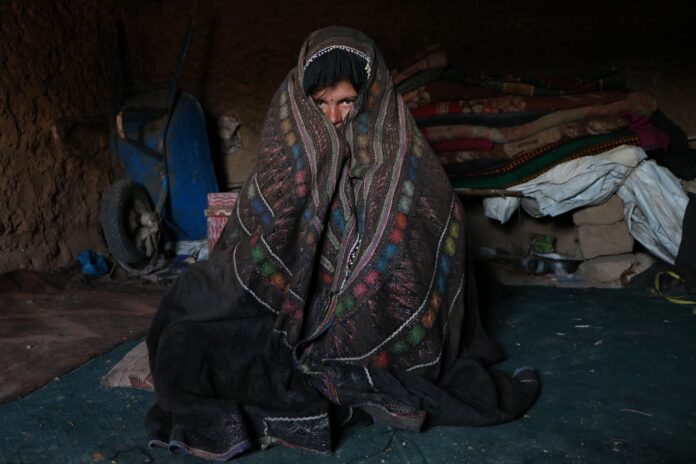 A person wrapped in a colorful blanket sitting in a dimly lit indoor space in Herat, Afghanistan. Photo by Morteza Rezaiy, pixel
